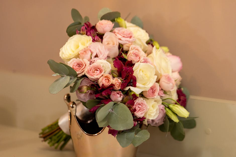 A fresh floral bouquet featuring a mix of pale pink and cream roses, complemented by deep burgundy flowers and green eucalyptus leaves, arranged in a compact, rounded style. The bouquet sits in a smal