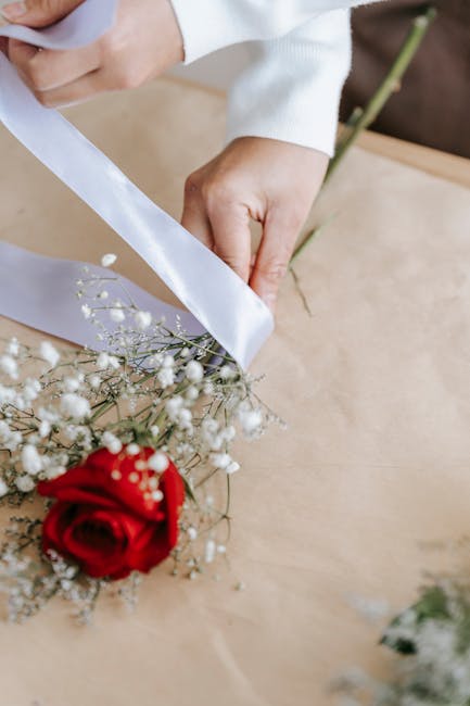 Close-up of a florist's hands, dressed in a white long-sleeve top, wrapping a fresh bouquet of flowers on a light wooden surface. The bouquet features a vibrant red rose, delicate white baby's breath,