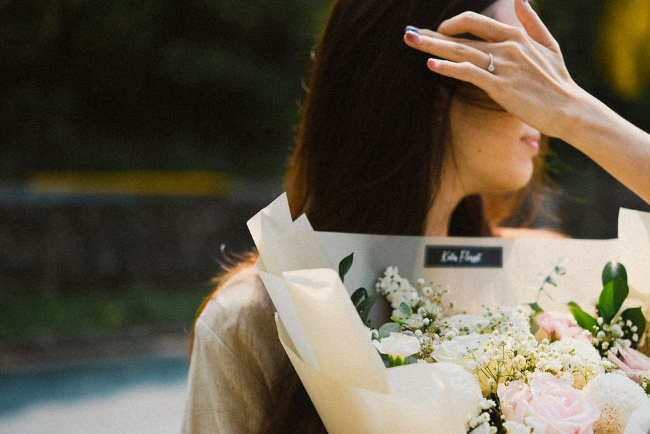 A woman with long, dark hair holds a large bouquet of fresh flowers, gently covering part of her face with her hand. The arrangement features a variety of blooms, including soft pink roses, clusters o