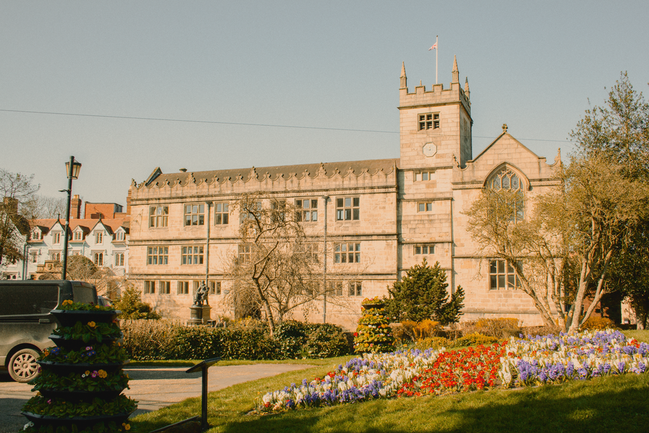 A vibrant flower bed featuring clusters of purple, red, yellow, and white flowers in full bloom, situated in front of an historic stone castle with a tower and battlements. The arrangement includes ne