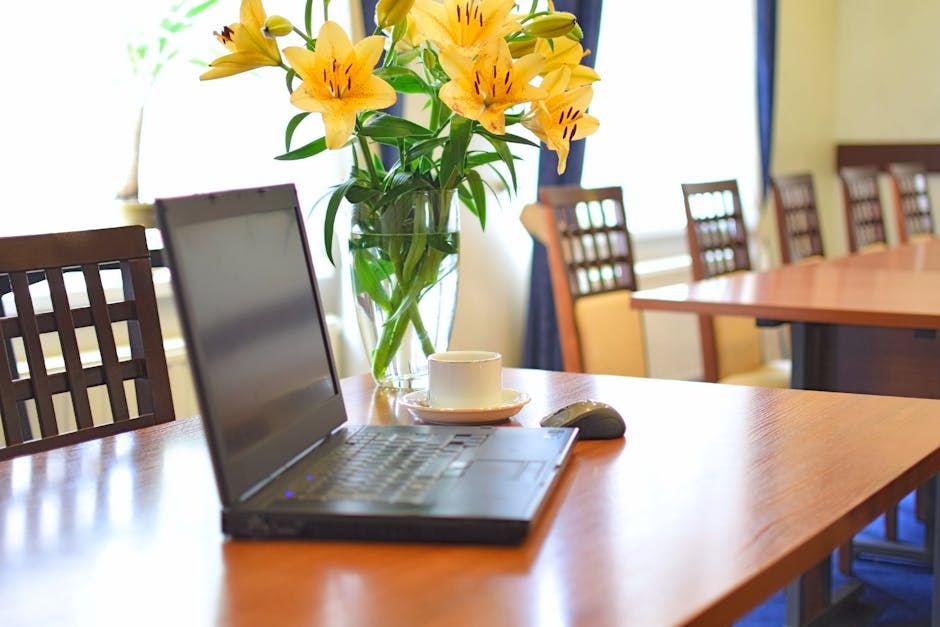 A clear glass vase filled with fresh yellow lilies, featuring large, open petals with dark streaks near the center, arranged on a polished wooden conference table. The bouquet is lush and vibrant, wit