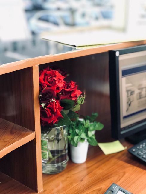 A floral arrangement of fresh red roses with dark crimson and bright red petals, accompanied by green foliage, is placed in a clear glass vase on a wooden office desk. The bouquet is partially obscure